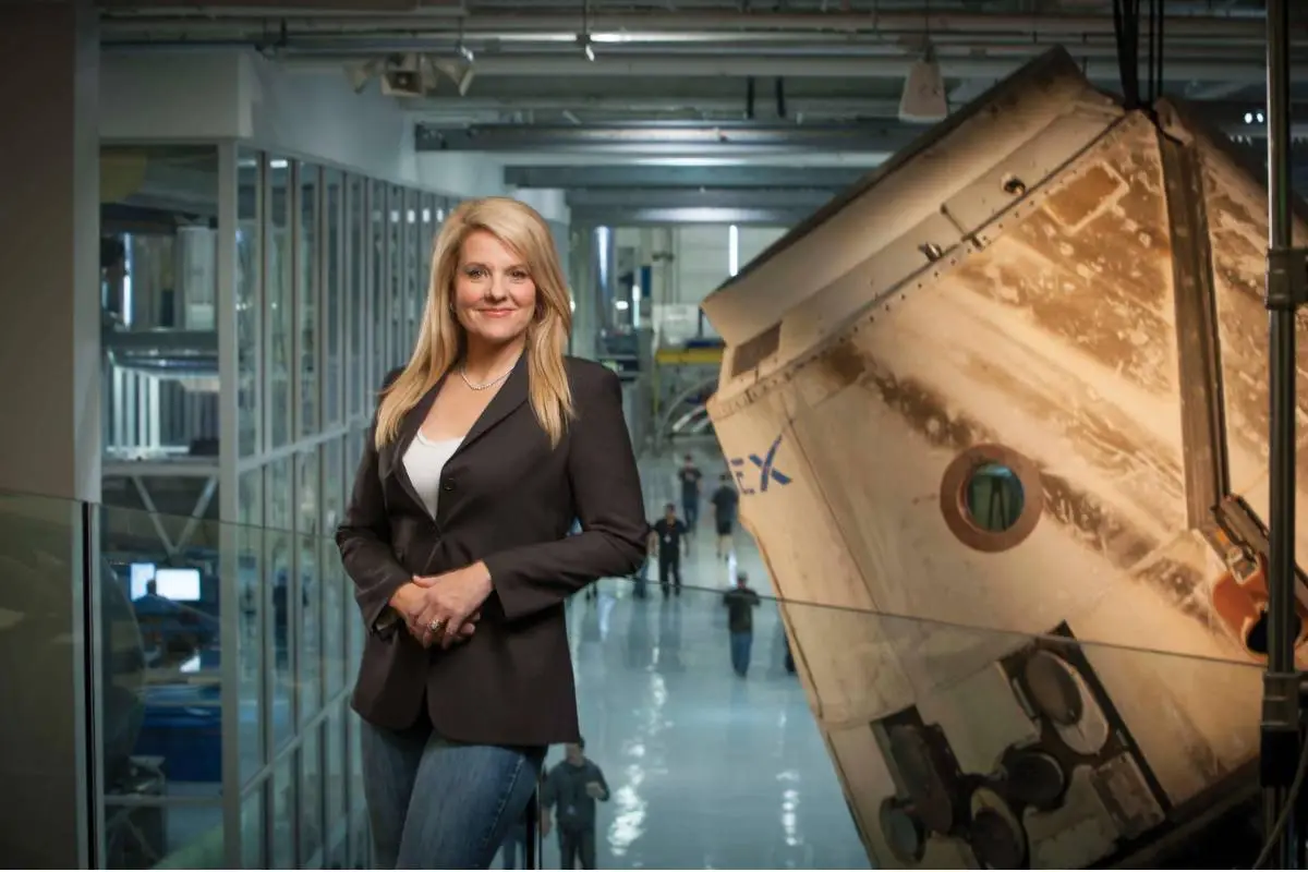 Gwynne Shotwell, presidente e COO da SpaceX, posando em um ambiente industrial moderno com uma cápsula espacial ao fundo, representando seu trabalho no desenvolvimento de tecnologia aeroespacial