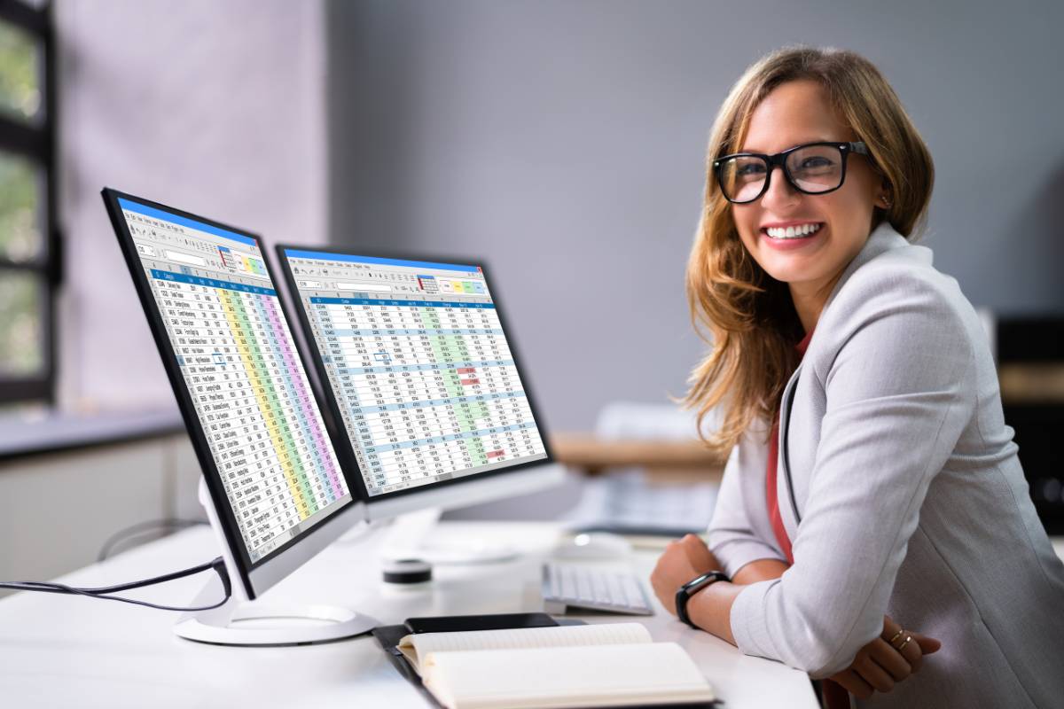 Mulher sorridente trabalhando com planilhas em dois monitores, em um escritório organizado e iluminado.